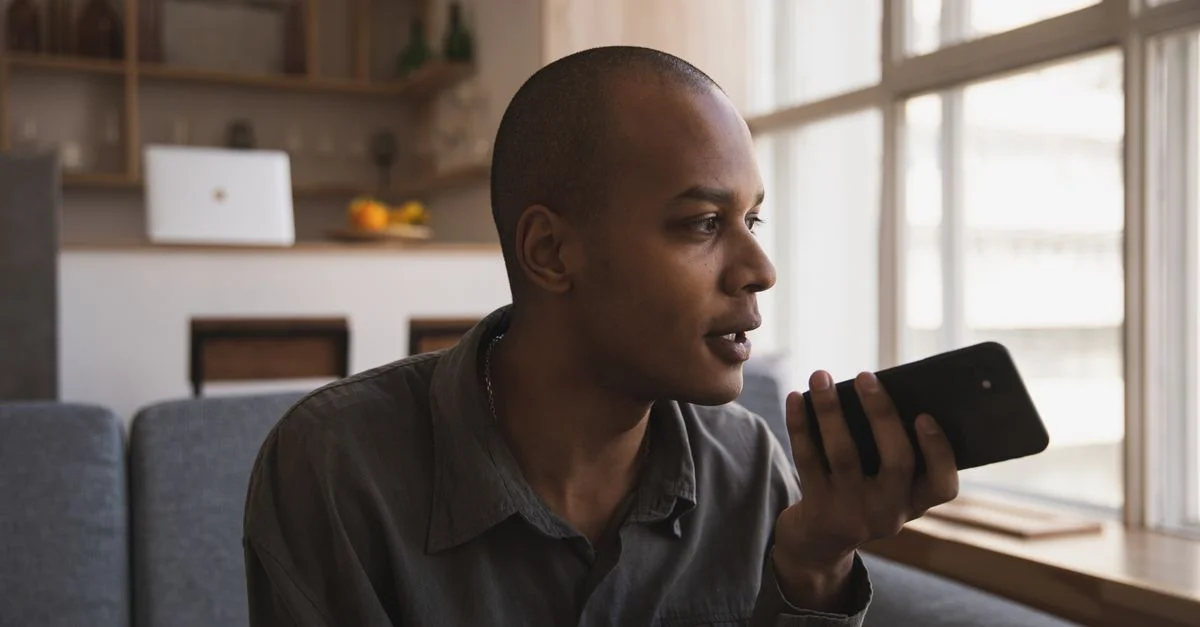 Homme tenant un smartphone et parlant à l'intérieur, assis sur un canapé près de la lumière de la fenêtre.