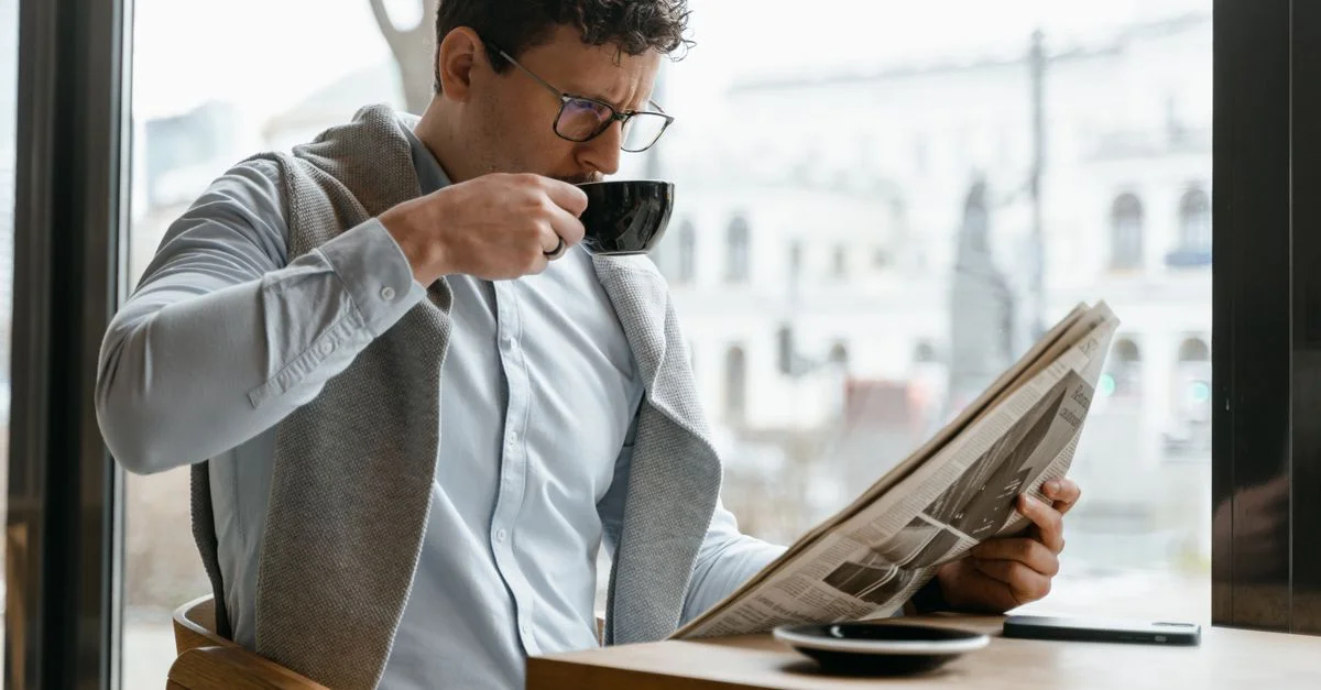 Homme adulte portant des lunettes, sirotant un café et lisant un journal dans une ambiance de café confortable.